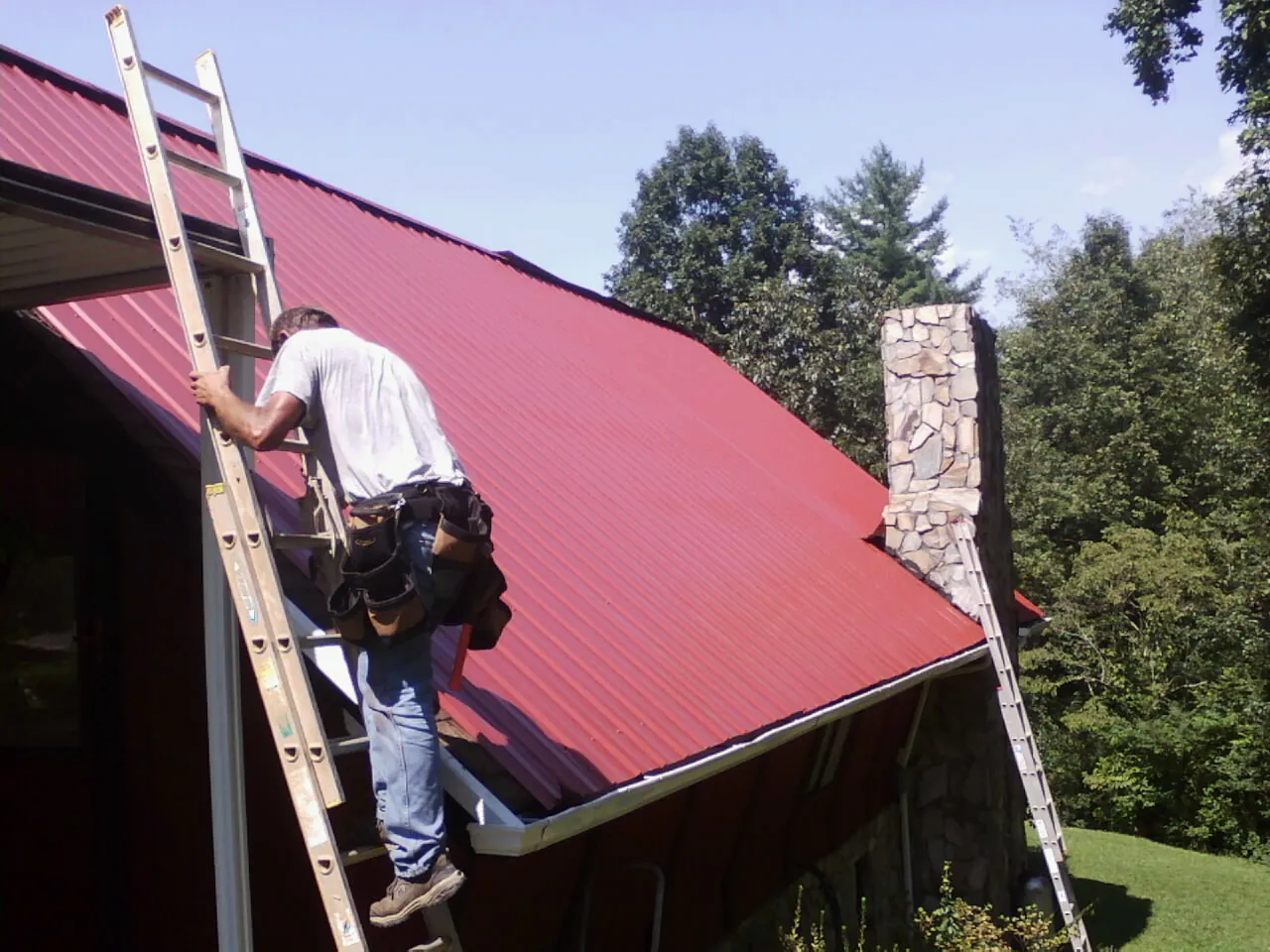 Roofer on a ladder beside a red metal roof and stone chimney — Soffit & Fascia Repair by Roofing Brothers in Brasstown